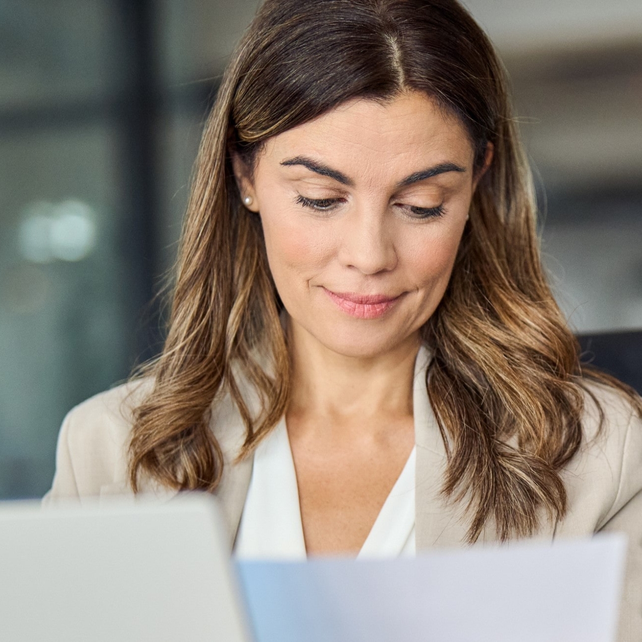 A white woman with mid-length light brown hair wearing a light coloured jacket sits at a laptop and looks at a sheet of paper